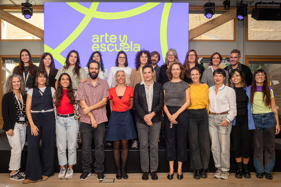 Grupo de participantes en la Jornada Arte y Escuela posando delante de una pantalla con la identidad gráfica del encuentro.